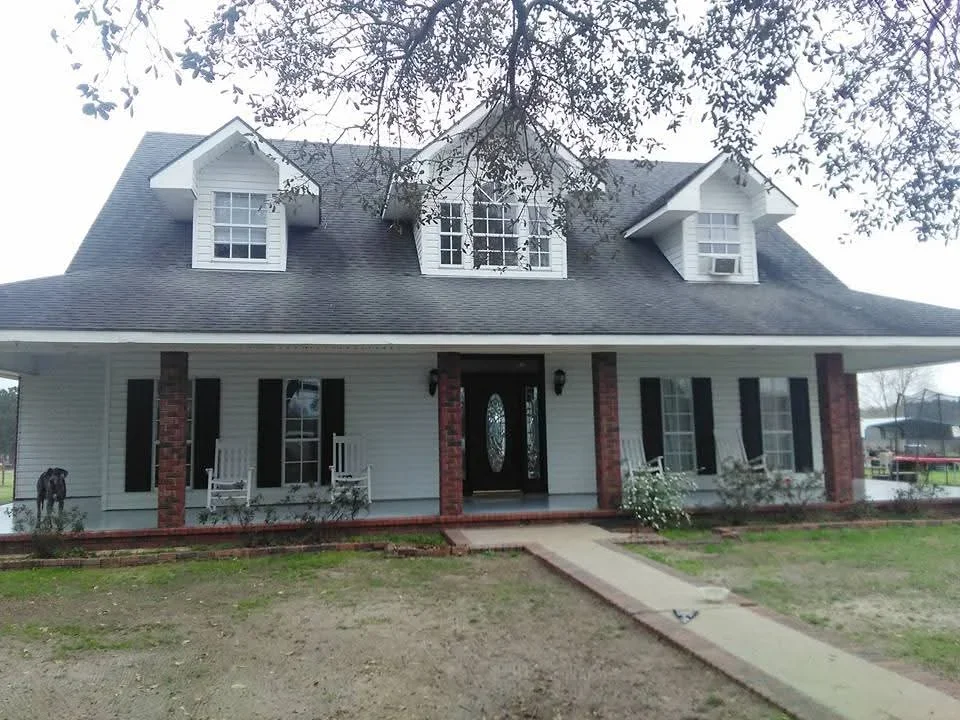 White farmhouse with black shutters, brick columns, and gray roof with dormers